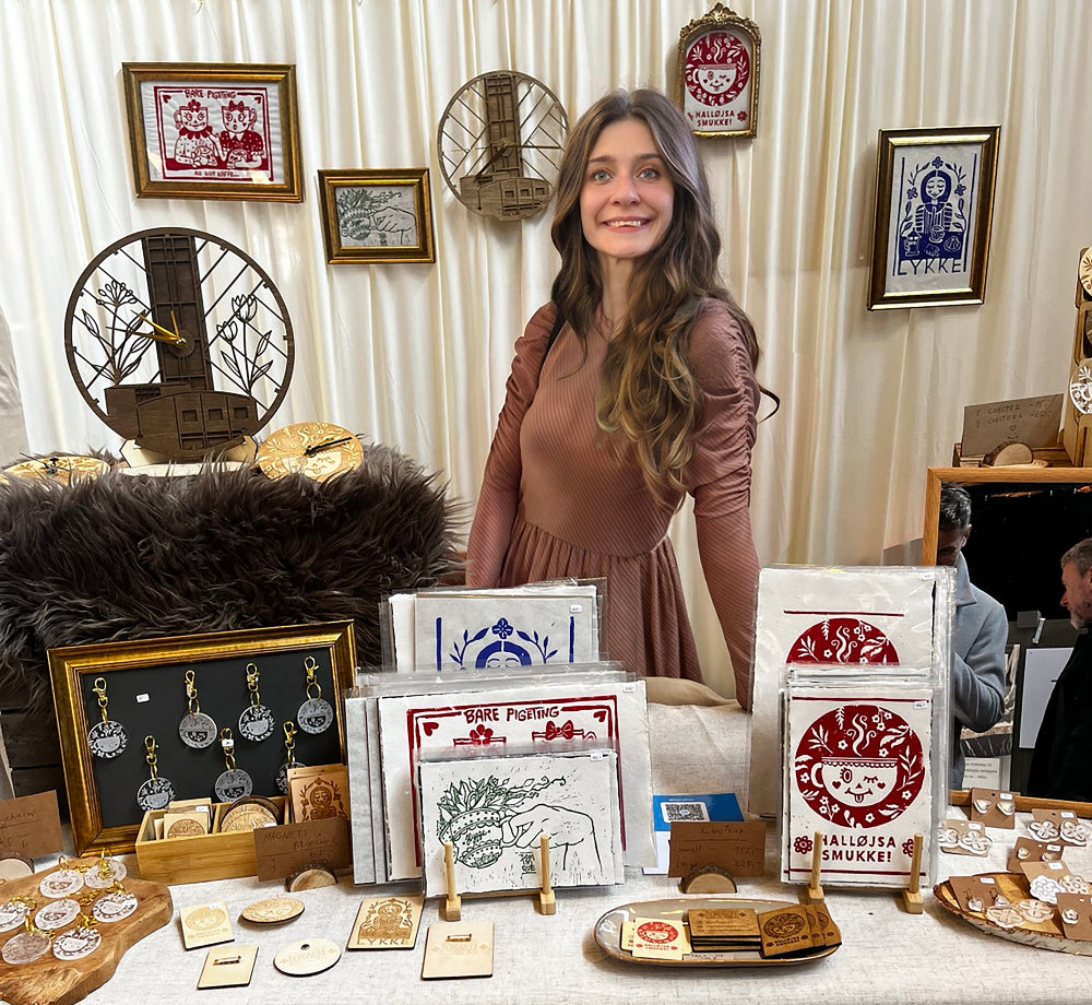 A woman stands behind a craft display table at an indoor market or fair, wearing a dusty rose long-sleeve dress. The table is covered with a white cloth and displays handmade items including framed art prints, wooden coasters, small wooden boxes, and jewelry.